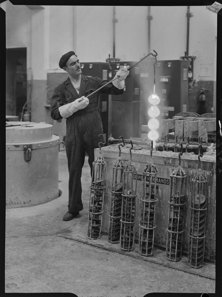 PANDO (1956) - Interior de la fábrica. Operario con mono de trabajo y guantes que sujeta una barra con metales incandescentes